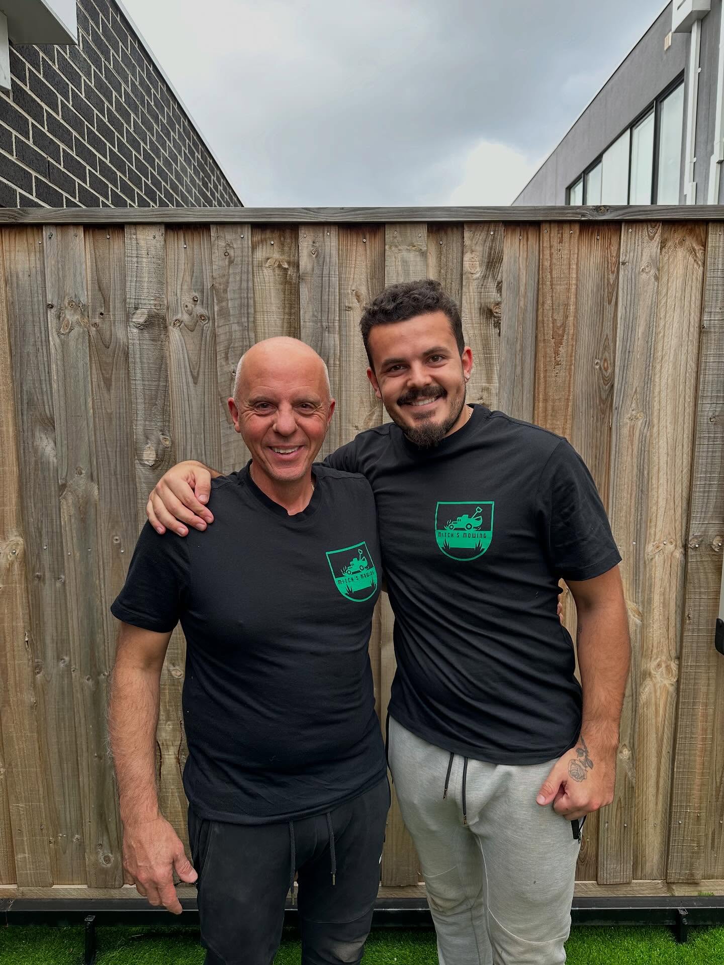 ac8 Two men in black logo T-shirts smiling in front of a timber fence