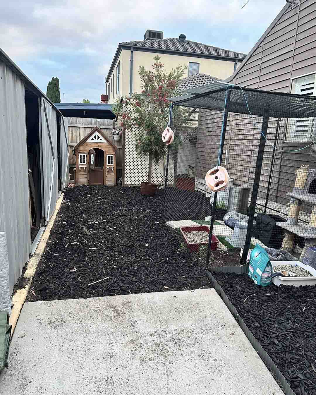 Fresh black mulch garden beds along a backyard path with a cubby house