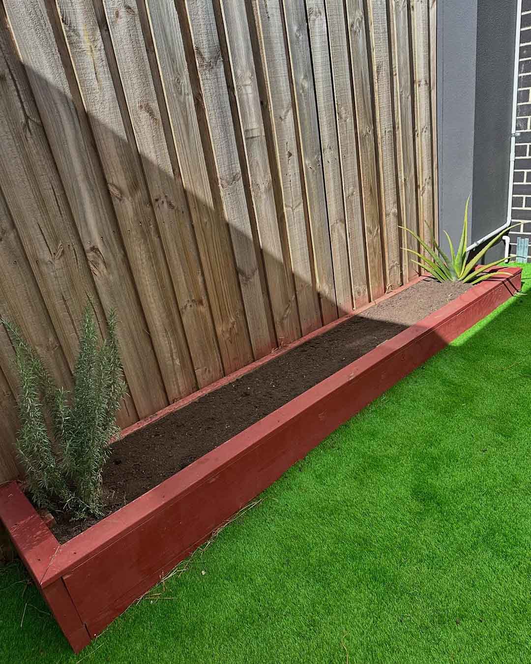 Red-edged raised garden bed with young plants along a fence above artificial turf